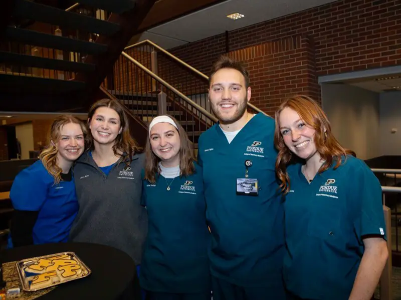 Five smiling people in Purdue-branded clothing pose together indoors.