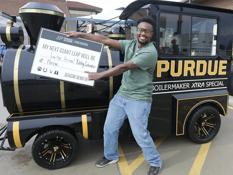 Smiling person holds a sign reading “My next giant leap will be at Purdue,” standing beside a Purdue-themed steam locomotive.