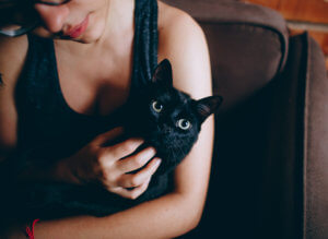 A person is holding a black cat, both looking at the camera with a cozy, intimate setting.