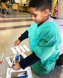 A child in green scrubs is kneeling on the floor, engaging with printed educational materials.