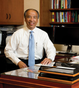 A professional portrait of a smiling man in formal attire sitting at a desk in an office setting.