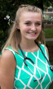 A professional-looking individual wearing a green patterned shirt and a stethoscope, smiling outdoors.