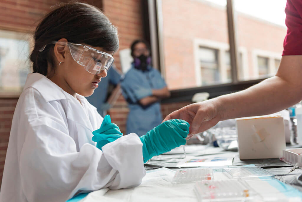 A young girl wearing gloves, a white coat, and safety goggles practices dropping liquid from a vial for testing as a hand reaches out to guide her.