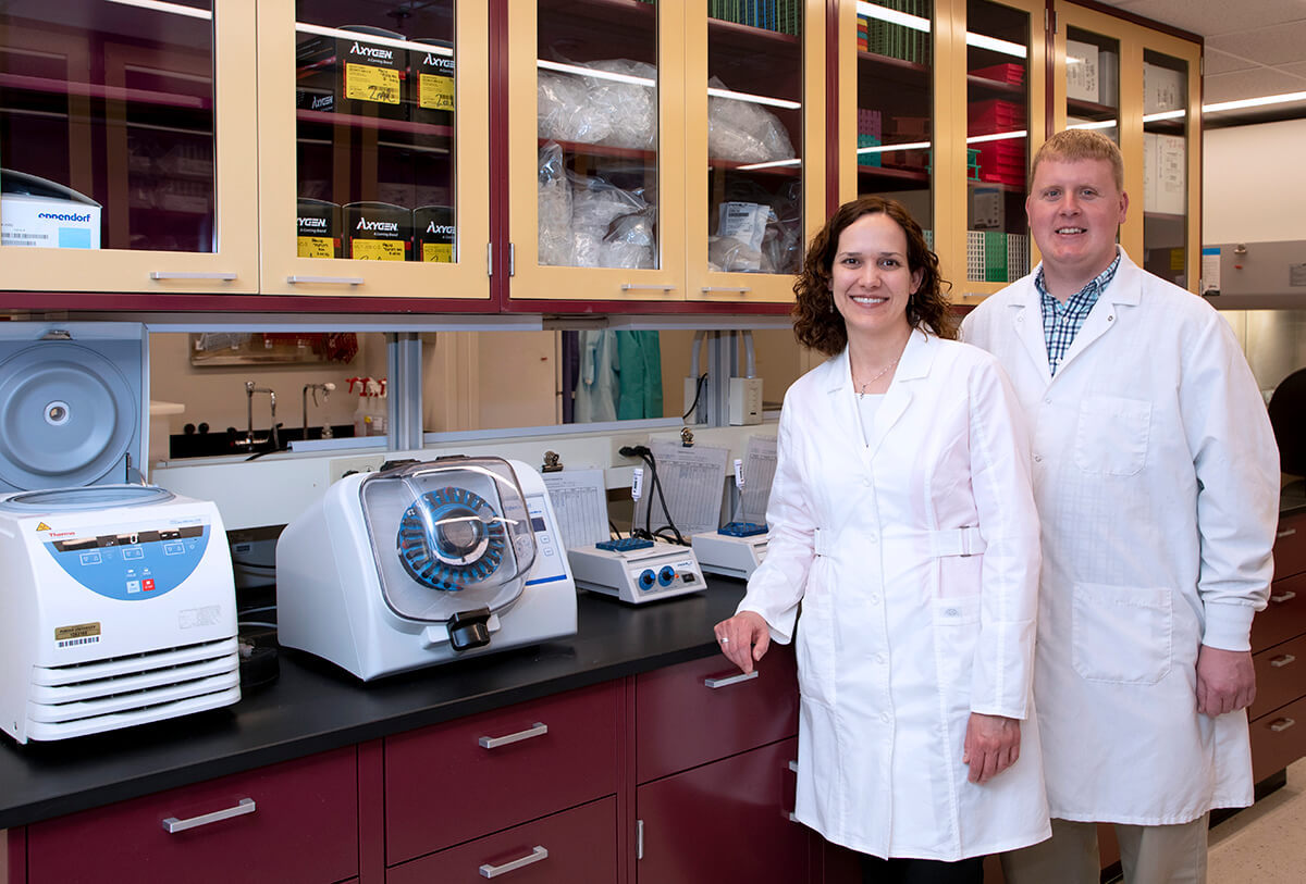 Dr. Hendrix stands in front of Dr. Bowen in front of a row of cabinets in a lab