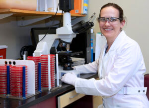 A laboratory setting featuring a smiling researcher in a white lab coat and safety glasses, positioned next to a microscope and laboratory equipment.