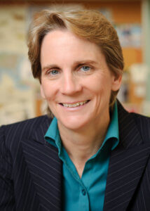 A headshot of a smiling individual with short, light brown hair, dressed in a striped blazer and blue shirt, set against a blurred background.