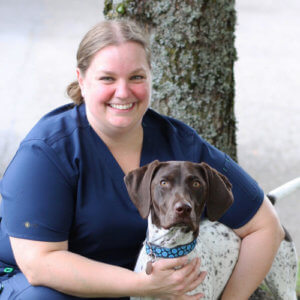 A smiling person in a blue uniform sits beside a brown and white dog against a tree.