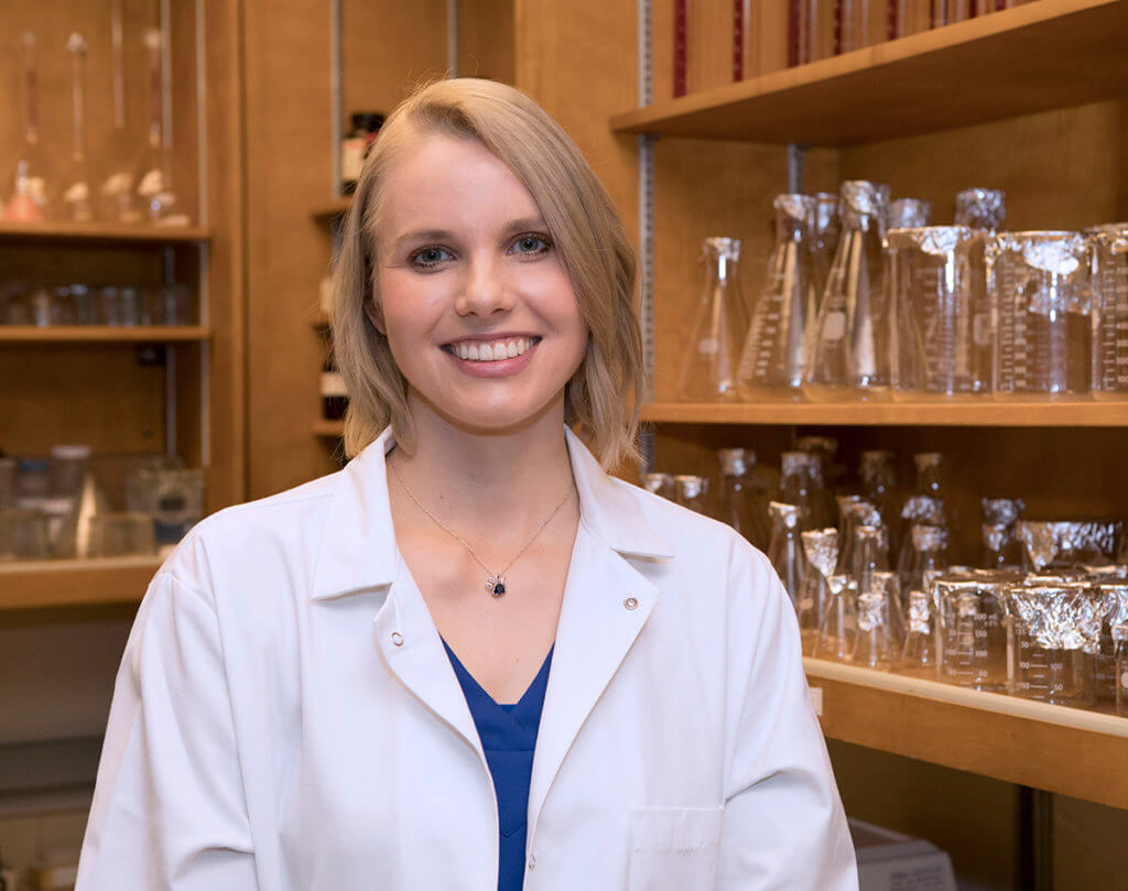 Alix stands in her laboratory wearing a white coat with shelves of beakers behind her