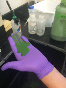 A gloved hand covered in green substance is shown, with laboratory equipment visible in the background.