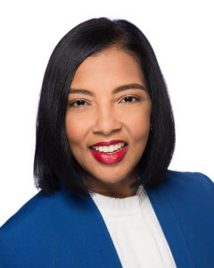 A headshot of a smiling person with shoulder-length black hair, wearing a blue blazer and red lipstick against a white background.