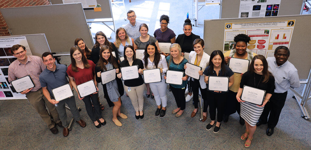Veterinary Research Scholars Summer Program participants hold up their certificates of recognition surrounded by program administrators during the research poster session in the library