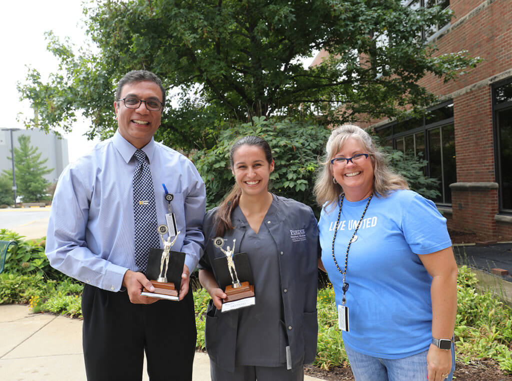 Dr. Narayanan and Christa hold up their corhnole tournament trophies joined by Andi outside Lynn Hall