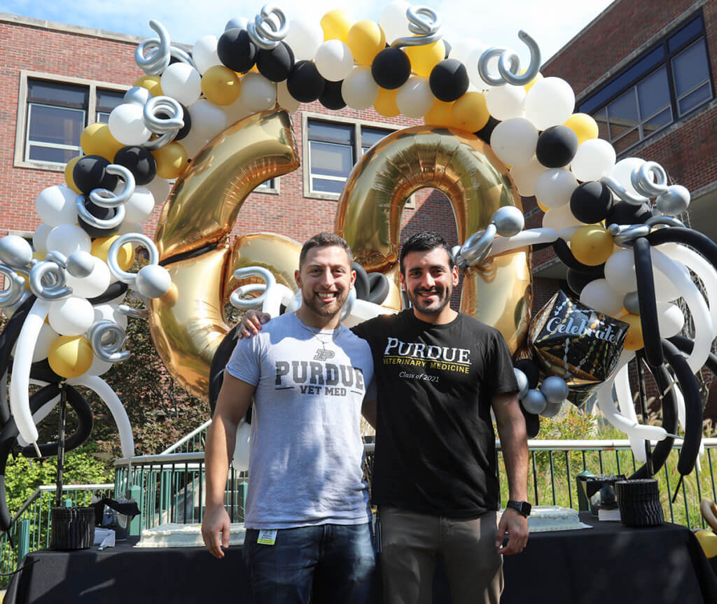 Matt and Javier stand in front of a cake table with a large balloon arch