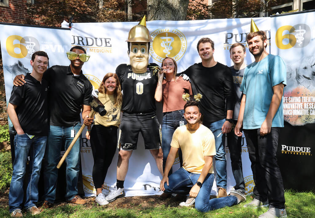 students gather around Purdue Pete against a photo backdrop donning party hats and accessories in the courtyard