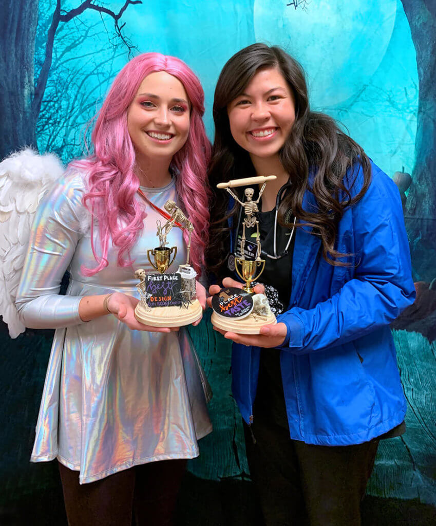 Brittany Rhodes dressed up as an angel and Bri Seiders hold their winning trophies against a spooky Halloween photo backdrop