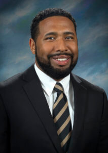 A professional headshot of a smiling man wearing a black suit and striped tie against a blurred background.