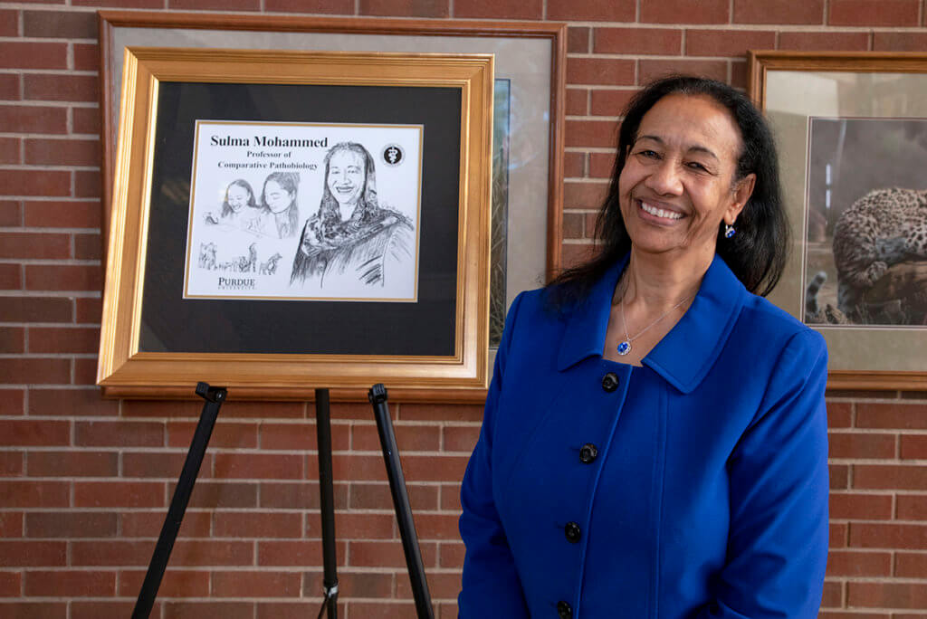 Dr. Mohammed stands next to a framed illustration of herself against a brickwall in the Lynn Hall Continuum Cafe