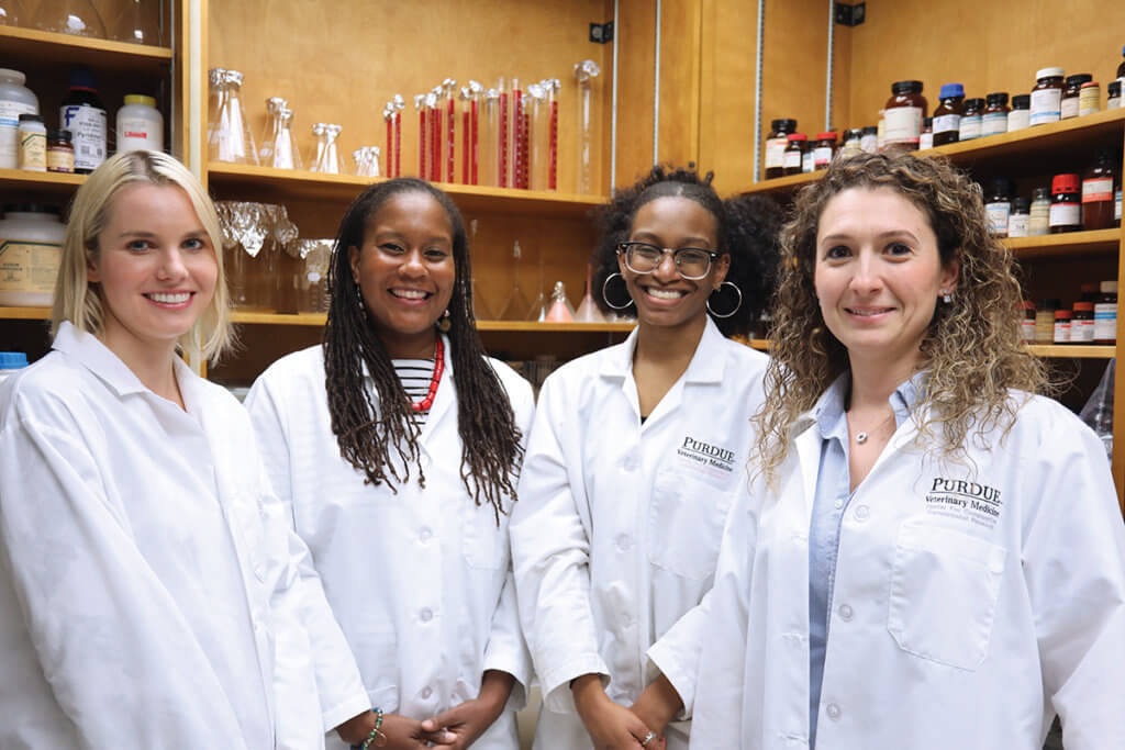 The lab team members stand together wearing white coats with laboratory equipment in the background