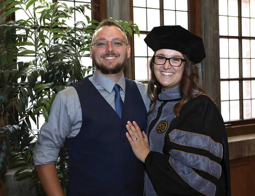 Dr. Taylor is dressed in her cap and gown standing next to her husband against a wall of windows in the Purdue Memorial Union