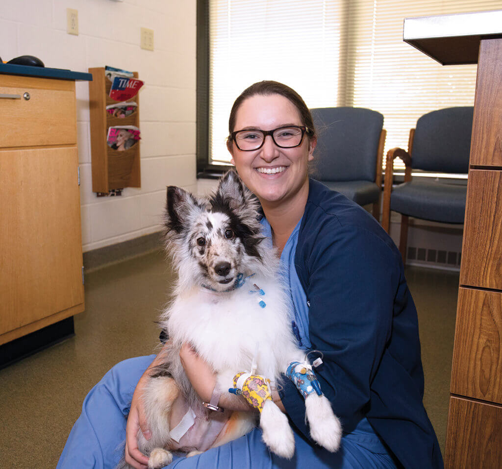 Taylor sits on the floor in a hospital exam room holding a black and white dog in her lap