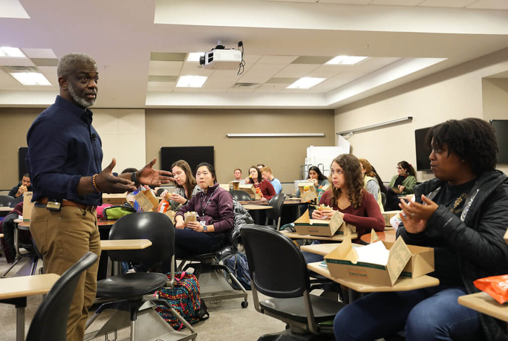 Ralph speaks to students as they eat lunch in a classroom