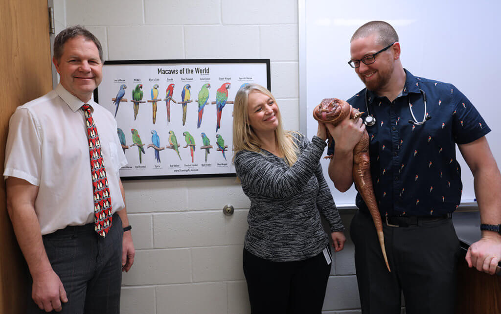 Joe holds Blaze while pets the reptile and Dr. Thompson stands to the side of Joe's framed drawing