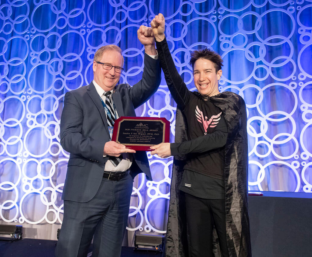 Sandy and Michael stand together on stage holding her award plaque with a fist in the air