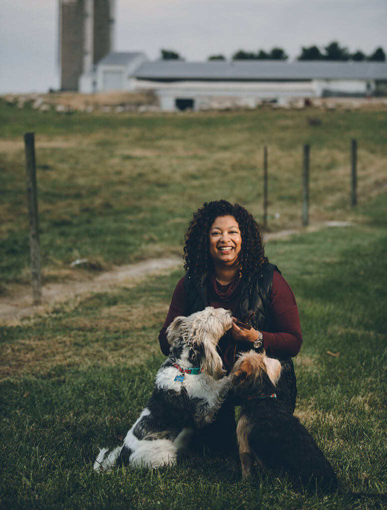 Candace is pictured kneeling in the grass with two dogs in front of her with a farm in the background