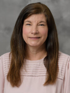 A portrait of a person smiling in a light-colored blouse against a gray background.