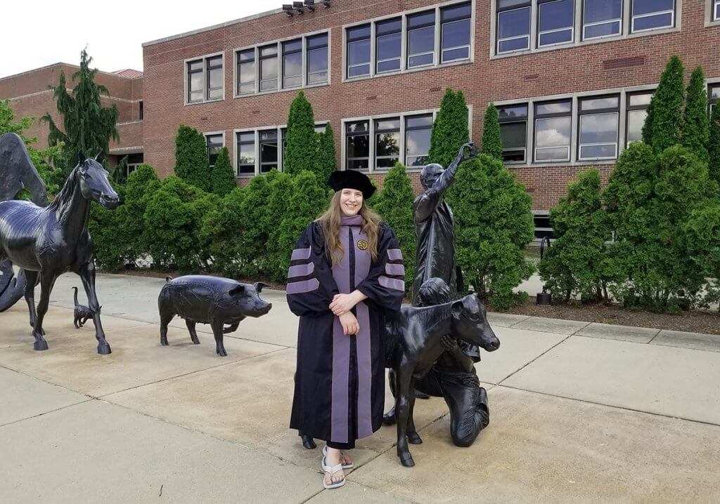 Colleeen stands in front of the Continuum sculpture wearing her graduation regalia