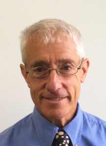 A headshot of an older man with gray hair and glasses, wearing a blue shirt and a patterned tie.