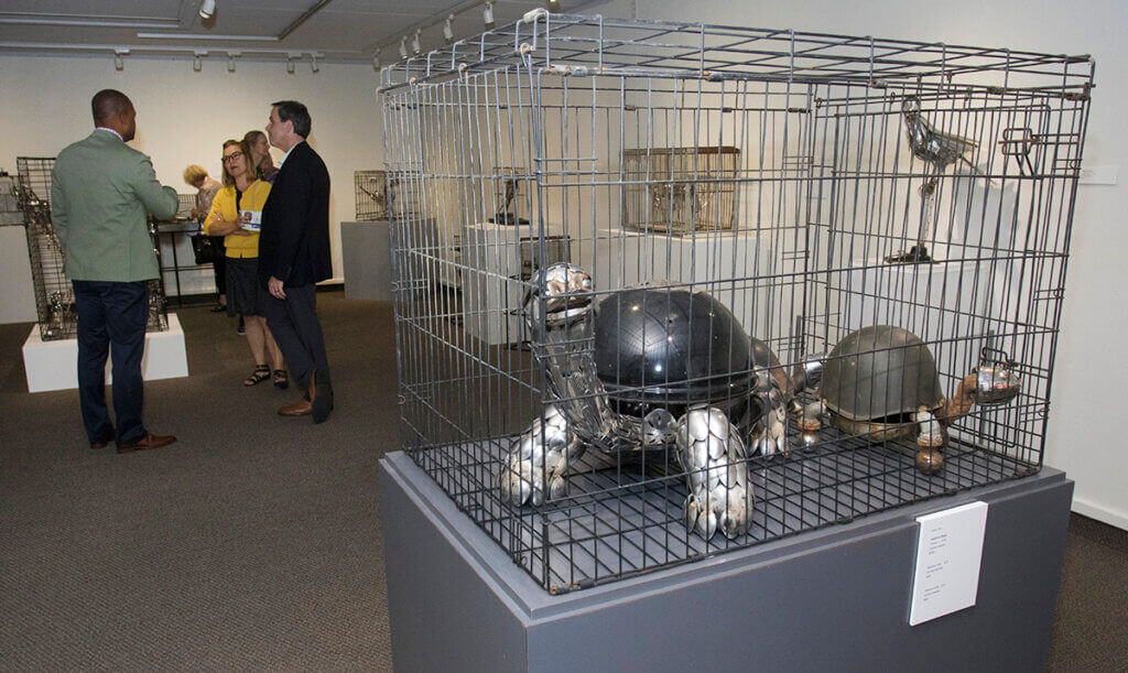 Metal sculpture of turtles in a cage is displayed in the museum with gallery attendees standing in the background