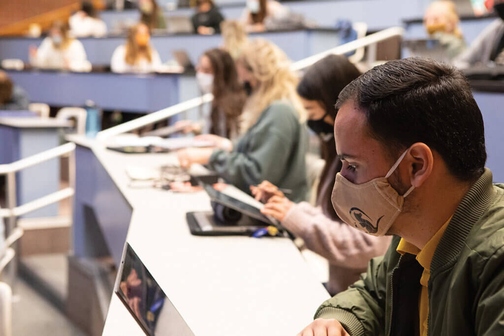 A student wears a face mask while looking at his laptop with fellow classmates seated around him in the background