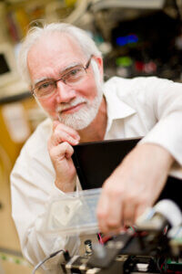 A portrait of an older man with glasses, smiling while interacting with electronic equipment in a laboratory setting.