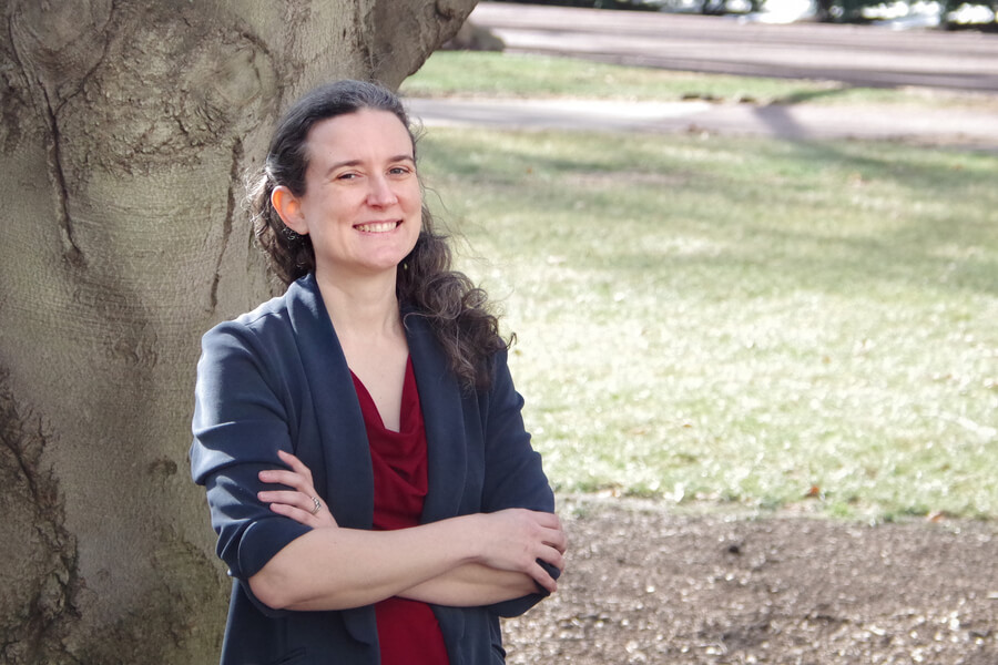 Kelly smiles with her arms crossed standing in front of a tree on a sunny day