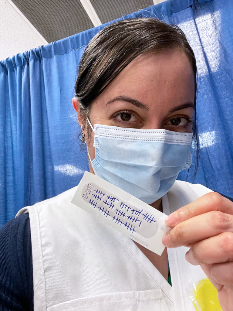 Dr. Malek wears a mask and volunteer vest as she holds up a bandaid marked with the number of vaccines she's administered