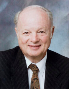 A headshot of a smiling older man wearing a dark suit and patterned tie.