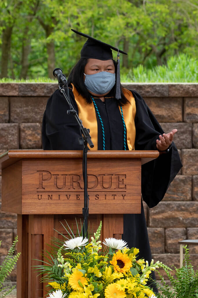 Bayli stands behind the podium wearing a cap and gown and a face mask