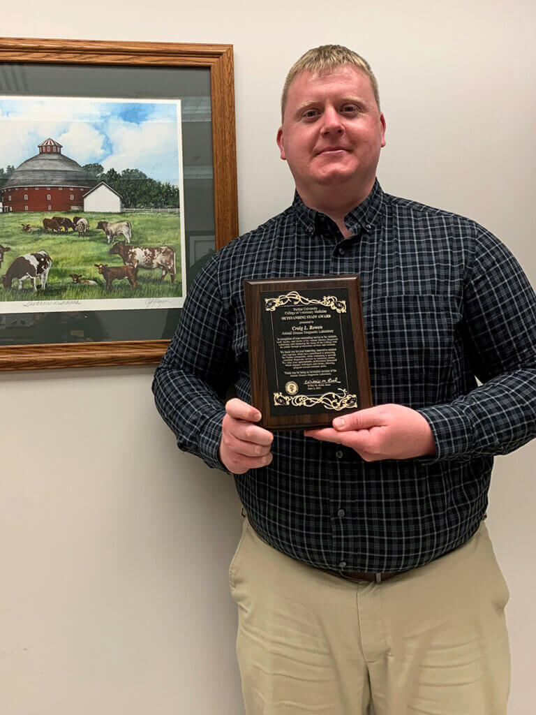 Craig stands next to a framed illustration of a farm scene holding up his award plaque and smiling