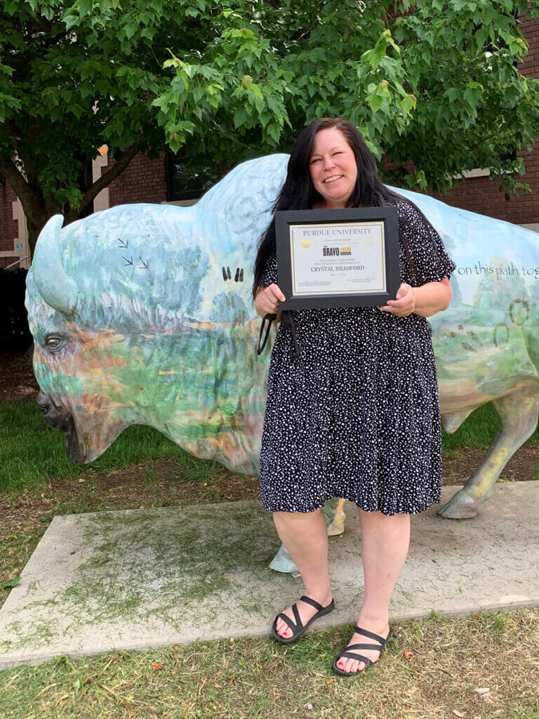 Crystal smiles standing in front of the bison statue in front of the Veterinary Pathobiology building holding up her Bravo Award certificate