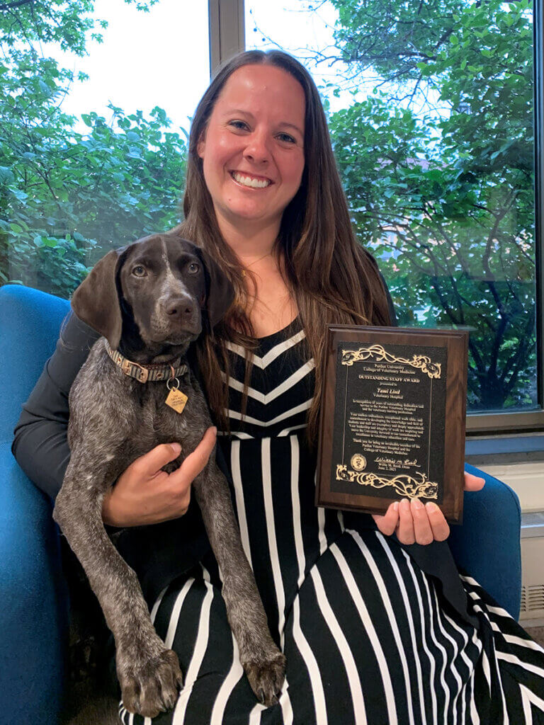 Tami smiles as she sits in a chair in the Small Animal Hospital reception holding a brown and gray speckled dog and her award plaque