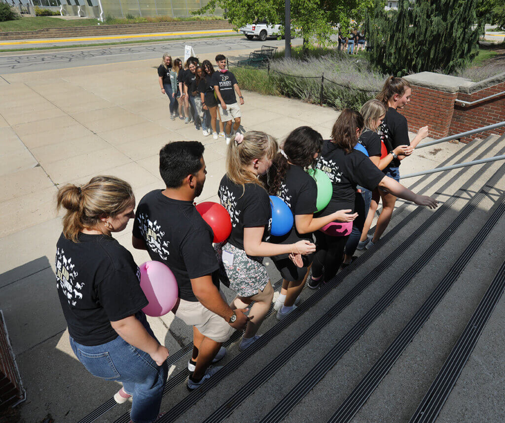 A group of students walk in a line on the stairs leading to Lynn Hall off Harrison Street with a balloon in between each person while another group walks toward them balancing balloons
