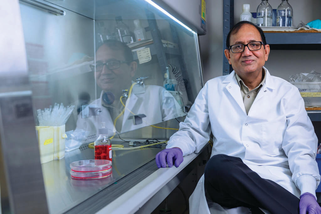 Dr. Mittal sits in his lab wearing a white coat and purple gloves smiling