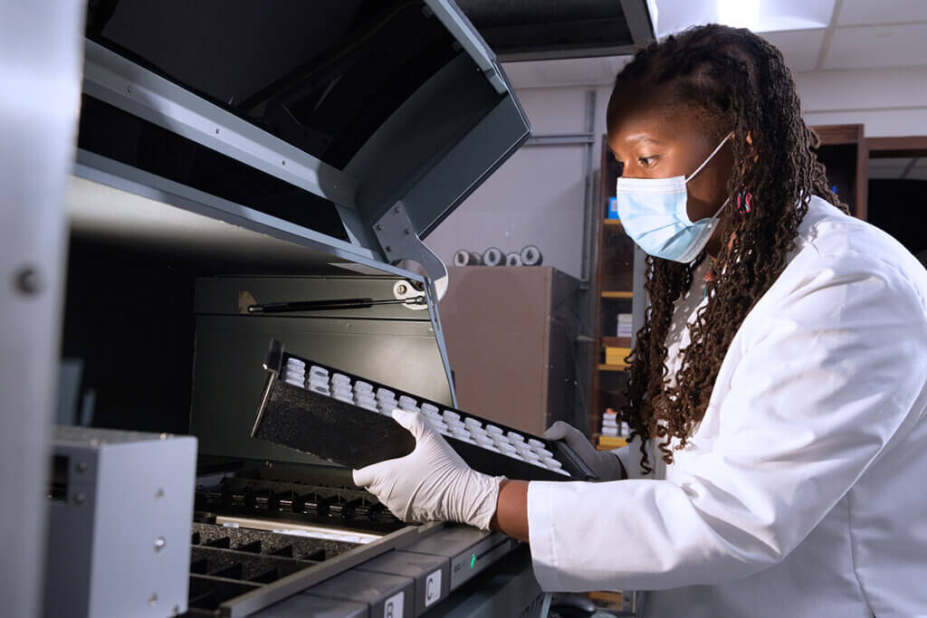 Dr. Lyle, wearing a white lab coat, gloves, and a mask, studies a tray of samples in her lab