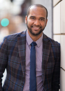 A smiling man in a plaid suit jacket stands against a wall, conveying a professional and approachable demeanor.