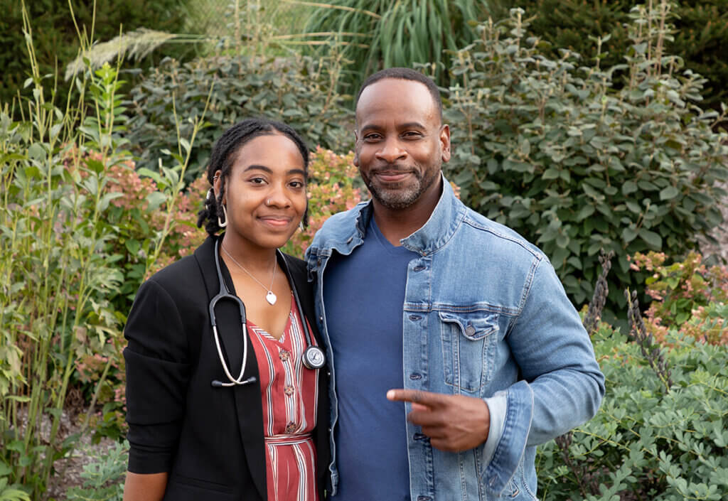 Myia smiles wearing her stethoscope standing next to her dad who is grinning and pointing to her with pride