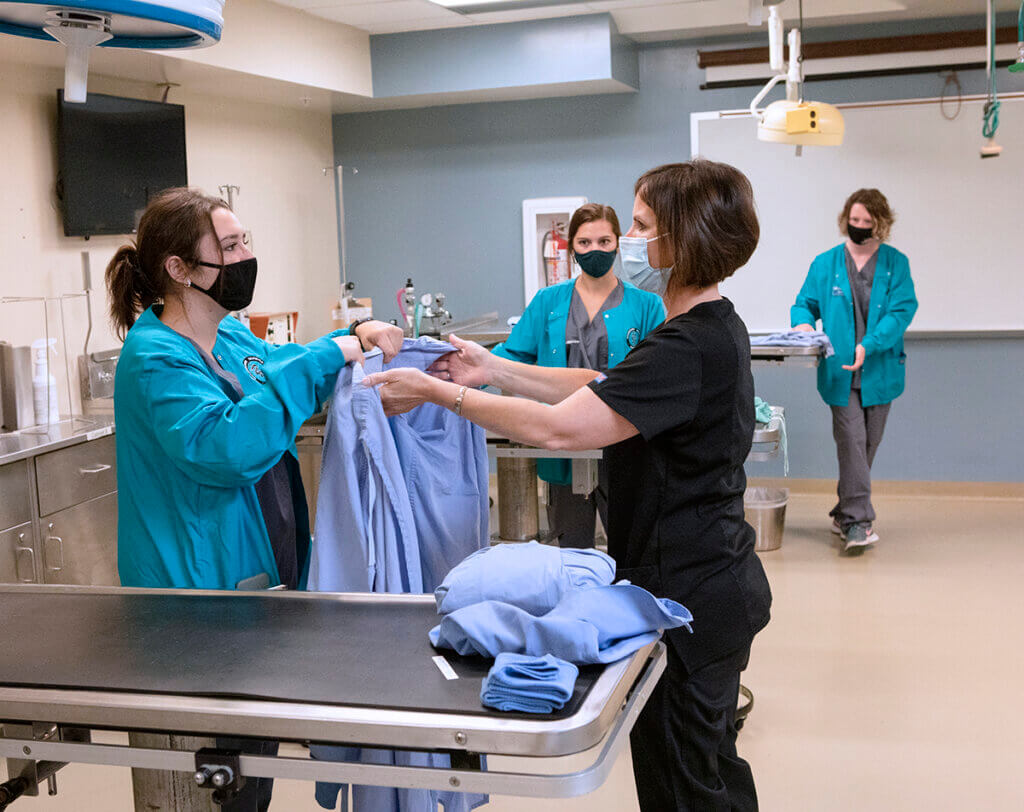 Cass and Kayleigh hold up a surgical gown beside an exam stable as two veterinary nursing students look on