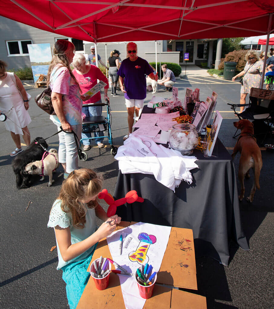 In the foreground, a girl sits decorating a bandana as Kelly chats with booth visitors in the background