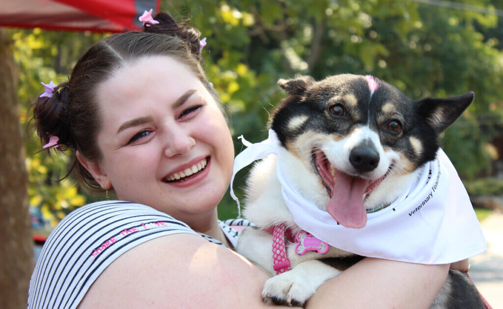 A woman smiles holding her small smiling black and white dog sporting a PVM bandana