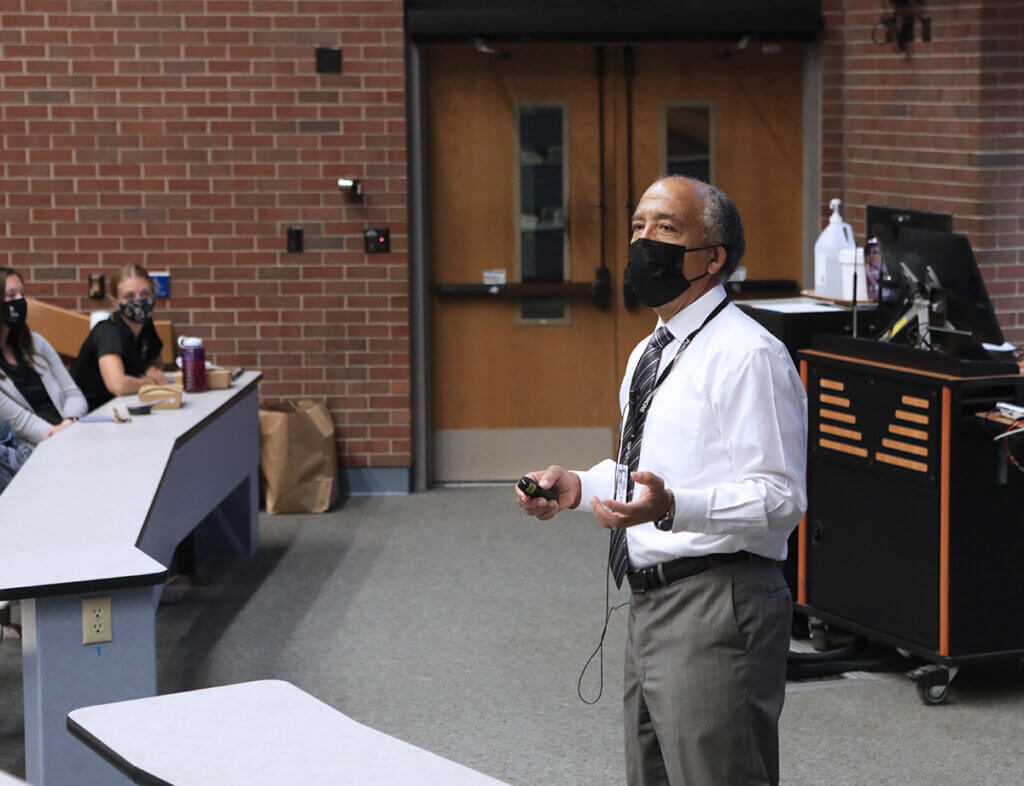 Dean Reed speaks to students from the front of the classroom wearing a mask and holding a presentation pointer